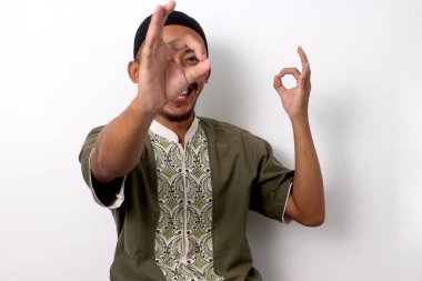 Koko ve peci 'deki mutlu Endonezyalı Müslüman adam, dürbün taklidi yaparken Ramazan ayındaki memnuniyetini dile getirirken, gözünün yanında eğlenceli bir jest yapıyor. Beyaz bir arkaplanda izole edilmiş