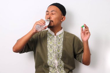 An Indonesian Muslim man in koko and peci breaks his Ramadan fast with a bottle of mineral water at sunset clipart