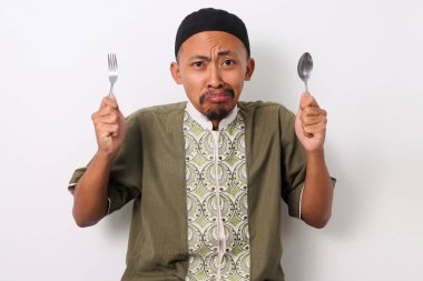 A hungry Indonesian Muslim man in koko and peci holds a fork and spoon, eagerly looking at the camera while anticipating the iftar meal to break his Ramadan fast. Isolated on a white background clipart