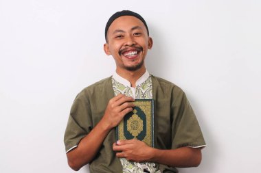 A joyous Indonesian Muslim man in koko and peci holds the Holy Quran to his chest, smiling warmly at the camera. Isolated on a white background clipart