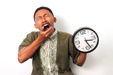 Sleepy Indonesian Muslim man in koko and peci stifles a yawn while checking a clock. He has just woken up to prepare for the suhoor meal before the Ramadan fast begins. Isolated on a white background clipart