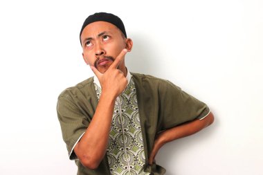 A pensive Indonesian Muslim man in koko and peci rests his hand on his chin in contemplation during Ramadan. Isolated on a white background clipart