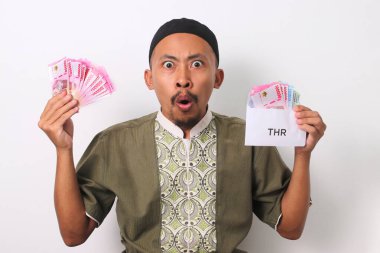 Şaşırmış bir ifadeyle şok olmuş Endonezyalı Müslüman bir adam, THR 'nin içinde para olan beyaz bir zarf tutuyor ve Dini Tatil Harçlığını temsil ediyor. Beyaz bir arkaplanda izole edilmiş