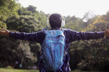 Sıradan kıyafetli, bereli, ekose gömlekli, sırt çantalı ve kulaklıklı genç bir Asyalı gülümseyerek kollarını açıyor. Doğa yürüyüşünde temiz havanın tadını çıkarıyor gibi görünüyor..