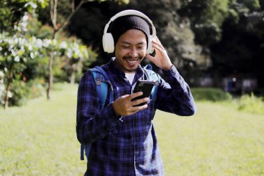 Günlük kıyafetli, bereli, ekose gömlekli, sırt çantalı ve kulaklıklı genç bir Asyalı. Telefonunda müzik arıyor. Görünüşe göre sabah yürüyüşü için en iyi çalma listesini ya da şarkıyı seçmiş..
