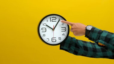 Parlak sarı bir arka planda duvar saatini tutan bir el. Zaman yönetimi, hatırlatıcılar veya geri sayım kavramını göstermek için ideal.