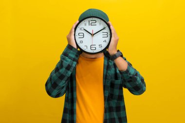 Şapka ve günlük gömlek giyen Asyalı bir adam, sarı bir arkaplana karşı durarak kafasını veya yüzünü örtmek için yaratıcı bir şekilde bir saat kullanır. Zaman yönetimi kavramı