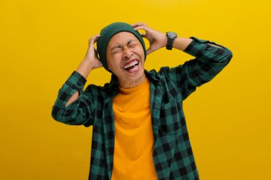 Çok neşeli, bereli ve rahat bir gömlek giyen Asyalı genç adam, harika haberler aldıktan sonra kendini şanslı hissederek mutluluk jesti olarak başını tutuyor. Sarı bir arkaplanda dikiliyorum.