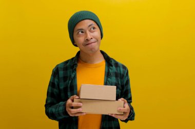 Bereli şapka ve günlük kıyafetler giyen düşünceli genç bir Asyalı paket paketleri tutuyor. Düşüncelere dalmış, boş fotokopi alanına bakıyor ve sarı bir zemine yaslanıyor.
