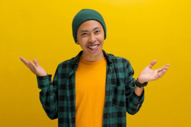 Şapka ve günlük giysiler giymiş gülümseyen genç bir Asyalı omuzlarını silkiyor, sarı bir arka plana karşı durarak bir seçenek veya karar sunuyor.