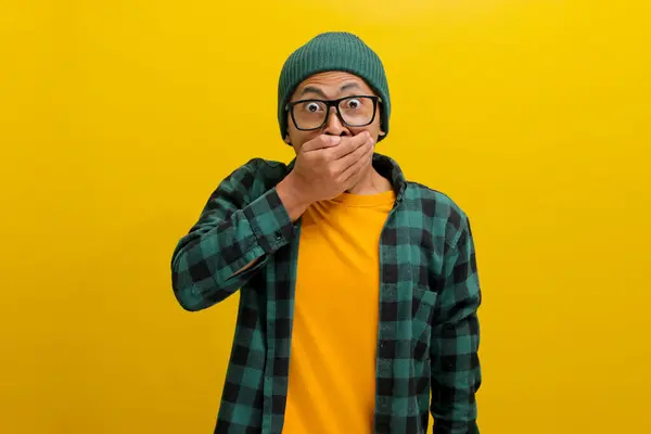 stock image Oops, surprised young Asian man, dressed in a casual shirt and beanie hat, covers his mouth, staring at the camera with an amazed expression while standing against yellow background.