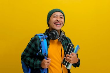 Bereli, sıradan kıyafetleri olan, sırt çantası ve kulaklık taşıyan, elinde bir yığın kitap tutan kendine güvenen Asyalı bir öğrenci. Sarı arka planda izole edilmiş. Akademik başarı, hazırlık, ya da öğrenme sevinci.