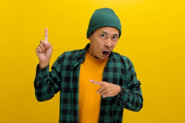 Şaşkın Asyalı bir adam, bereli bir şapka ve günlük bir gömlek giymiş, boş bir yeri işaret ediyor, muhtemelen bir ürünü tanıtıyor ya da inanılmaz bir teklif hakkında bilgi paylaşıyor, sarı arka planda izole edilmiş..
