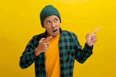Şaşkın Asyalı bir adam, bereli bir şapka ve günlük bir gömlek giymiş, boş bir yeri işaret ediyor, muhtemelen bir ürünü tanıtıyor ya da inanılmaz bir teklif hakkında bilgi paylaşıyor, sarı arka planda izole edilmiş..