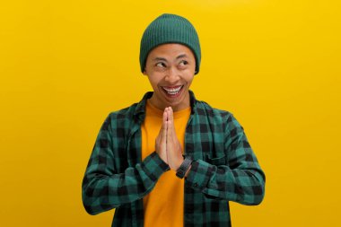 Asyalı heyecanlı genç bir adam, bereli bir şapka ve günlük bir gömlek giyerek, sarı bir arka planda dururken sevinç ve zaferi bir yumruk pompasıyla ya da EVET hareketiyle kutluyor.