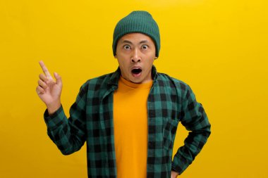 Şaşkın Asyalı bir adam, bereli bir şapka ve günlük bir gömlek giymiş, boş bir yeri işaret ediyor, muhtemelen bir ürünü tanıtıyor ya da inanılmaz bir teklif hakkında bilgi paylaşıyor, sarı arka planda izole edilmiş..