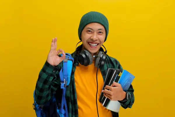 stock image Confident Asian student in beanie and casual clothes, carrying backpack and headphones, Holding a stack of books. Isolated on yellow background. Academic success, preparation, or the joy of learning.