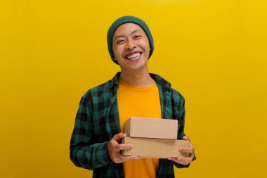 Şapka ve günlük gömlek giymiş gülümseyen Asyalı bir adam, sarı bir zemin üzerinde kameraya neşe içinde bakarken bir paket kutusu yığını tutuyor.