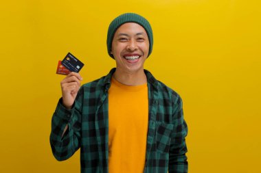 Şapka ve günlük gömlek giymiş neşeli, gülümseyen Asyalı genç bir adam, bir kredi kartı tutuyor, başarısını kutluyor, sarı bir arka plana karşı durarak