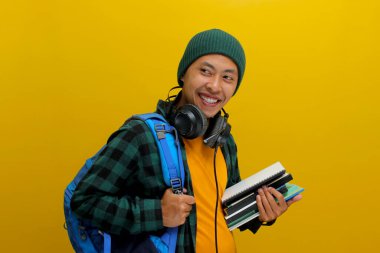 Bereli, sıradan kıyafetleri olan, sırt çantası ve kulaklık taşıyan, elinde bir yığın kitap tutan kendine güvenen Asyalı bir öğrenci. Sarı arka planda izole edilmiş. Akademik başarı, hazırlık, ya da öğrenme sevinci.