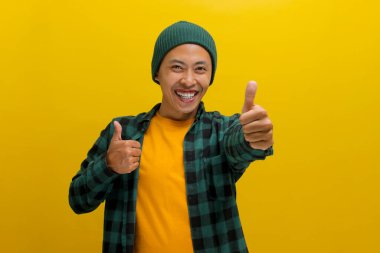 Sıradan giyinmiş ve bereli bir şapka takmış gülümseyen Asyalı genç bir adam, sarı arka planda dururken olumlu veya iyi bir eleştiriyi işaret eden bir jest yapıyor.