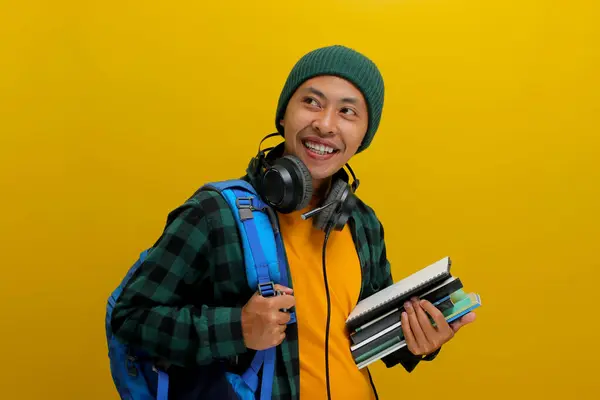 stock image Confident Asian student in beanie and casual clothes, carrying backpack and headphones, Holding a stack of books. Isolated on yellow background. Academic success, preparation, or the joy of learning.