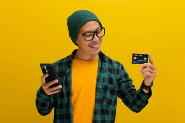 Şapka ve günlük giysiler giymiş genç bir Asyalı, sarı arka planda dururken telefonunu ve kredi kartını online alışveriş için kullanıyor.