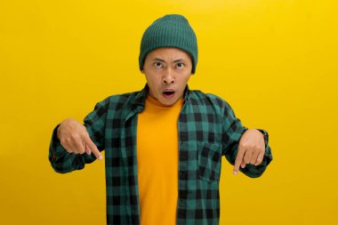 Şaşkın Asyalı bir adam, bereli bir şapka ve günlük bir gömlek giymiş, boş bir yeri işaret ediyor, muhtemelen bir ürünü tanıtıyor ya da inanılmaz bir teklif hakkında bilgi paylaşıyor, sarı arka planda izole edilmiş..