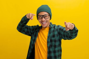 Sensin. Şapka ve günlük tişört giymiş genç Asyalı adam kameraya parmağını doğrultup gülümsüyor, insanları seçiyor, davet ediyor, adam topluyor, sarı arka planda dikiliyor.