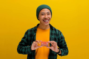 Şapka ve günlük gömlek giyen mutlu bir Asyalı genç adam, elinde bir kredi kartı, online alışveriş ödemesi için bir çözüm gösterirken heyecanlanıyor.