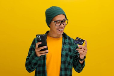 Şapka ve günlük giysiler giymiş genç bir Asyalı, sarı arka planda dururken telefonunu ve kredi kartını online alışveriş için kullanıyor.