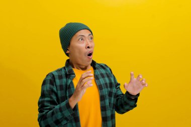 Asyalı heyecanlı genç bir adam, bereli bir şapka ve gündelik gömlek giymiş, yüksek sesle haykırıyor, elleri sarı bir arka planda dururken şaşkınlıkla hareket ediyor