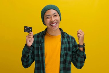 Şapka ve günlük gömlek giymiş neşeli, gülümseyen Asyalı genç bir adam, bir kredi kartı tutuyor, başarısını kutluyor, sarı bir arka plana karşı durarak