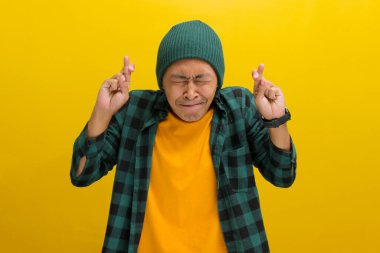 Şapka ve günlük gömlek giymiş, umut dolu genç Asyalı bir adam, sarı bir arka plana karşı durarak parmaklarını çaprazlarken bir dilek diler.