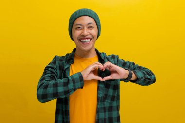 Çekici Asyalı bir adam, bereli bir şapka ve gündelik bir gömlek giyer. Elleriyle romantik bir kalp jesti yaratırken mutluluk saçar.