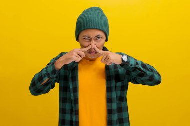 Şapka ve günlük gömlek giyen genç Asyalı adam, muhtemelen sarı arka planda dururken kötü kokudan dolayı parmaklarıyla burnunu çimdikliyor..