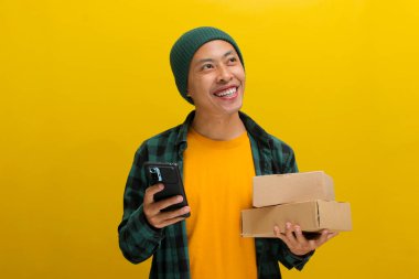 Şapka takan genç bir Asyalı adam, birden fazla iş yapıyor. Sarı bir arka plana karşı elinde karton bir kutu tutarken telefonunu kullanıyor.