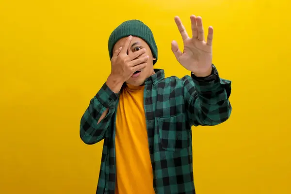 stock image Fearful young Asian man, dressed in a beanie hat and casual shirt, covers his eyes, possibly shielding himself from the frightful or overwhelming scenes while watching horror movies during Halloween