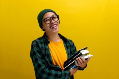 Genç bir Asyalı öğrenci, gözlüklü, bereli, sıradan elbiseli, sarı arka planda duruyor, göğsüne kitap dayamış..