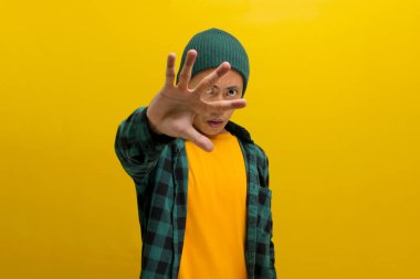 Ciddi bir Asyalı genç adam, bereli bir şapka ve günlük giysiler içinde, sıkı bir DUR hareketi yapıyor. Elleriyle, sarı arka plana karşı durarak bir ret, yasaklama ve uyarı işareti veriyor.