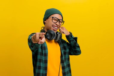 Hevesli Asyalı genç bir adam, bereli bir şapka ve sıradan bir gömlek giyer, enerjik bir şekilde kameraya doğru bakarken sarı arka planda dikilirken beni arar..