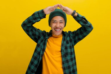 Şapkalı ve günlük kıyafetleri olan mutlu Asyalı genç adam başının üstünde el hareketi yapıyor. Sarı arka planda ayakta dururken çatıyı ya da evi simgeliyor.