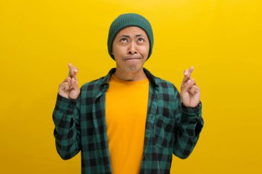 Şapka ve günlük gömlek giymiş, umut dolu genç Asyalı bir adam, sarı bir arka plana karşı durarak parmaklarını çaprazlarken bir dilek diler.