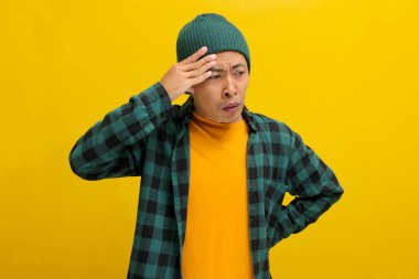 Şapka ve günlük gömlek giymiş genç Asyalı bir adam başını tutuyor. Bu da sarı arka planda izole edilmiş bir baş ağrısı yaşadığını gösteriyor. Sağlık hizmetleri ve tıbbi kavramlar