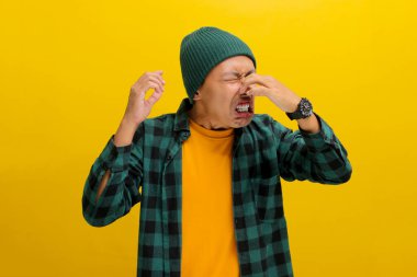 Şapka ve günlük gömlek giyen genç Asyalı adam, muhtemelen sarı arka planda dururken kötü kokudan dolayı parmaklarıyla burnunu çimdikliyor..