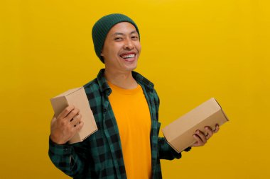 Şapka ve günlük gömlek giymiş gülümseyen Asyalı bir adam, sarı bir zemin üzerinde kameraya neşe içinde bakarken bir paket kutusu yığını tutuyor.