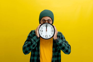 Şapka ve günlük gömlek giyen Asyalı bir adam, yüzünün önünde bir saat tutuyor, şaşırmış ve komik bir ifadeyle kameraya bakıyor, sarı bir arkaplan karşısında dururken