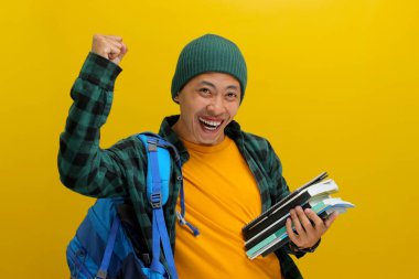 Asyalı bir öğrenci, bereli bir şapka ve günlük bir gömlek giyer ve sırt çantası taşır. Elinde kitap tutarken kolunu kaldırır ve eğitim yolculuğundaki başarısını kutlar.
