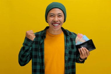 Heyecanlı Asyalı adam, bereli bir şapka takıyor ve sıradan kıyafetler giyiyor. Yumruğunu kaldırıyor. Başarını kutlarken sarı arka planda izole edilmiş parayı tutuyor..