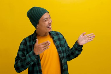 Asyalı heyecanlı genç bir adam, bereli bir şapka ve gündelik bir gömlek giyer, kollarını boş bir alana doğru uzatır, sarı bir arka plana karşı durarak birisini kendisine katılması için davet eder.